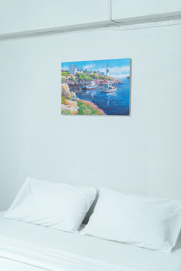 A bedroom with a bed, white bedding, and a painting of a coastal scene on the wall. - Nicha Residence Hotel (Bangkok)