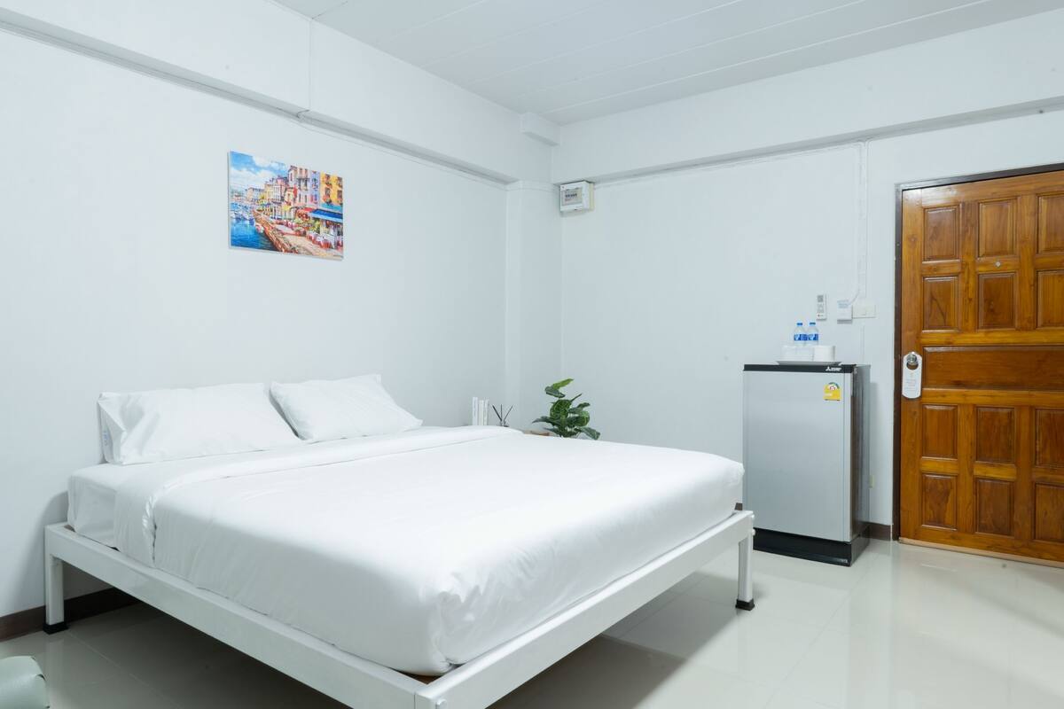 A hotel room with a single bed, a small refrigerator, a wooden door, and a painting on the wall.