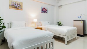 A hotel room with two single beds, a bedside table with a lamp, a small desk with a refrigerator, and two paintings on the wall.