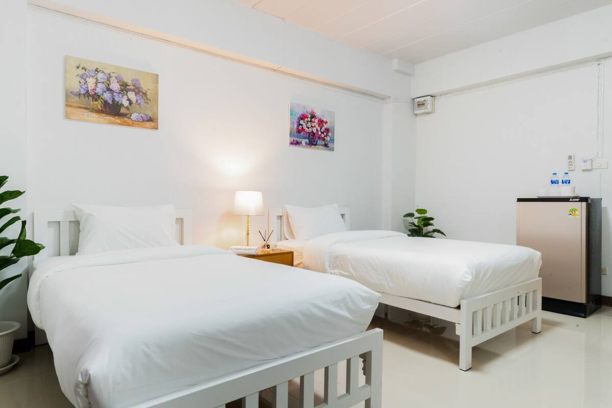 A hotel room with two single beds, a bedside table with a lamp, a small desk with a refrigerator, and two paintings on the wall.