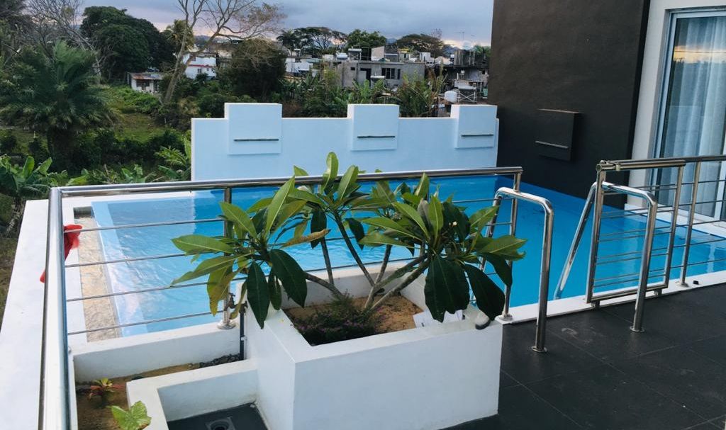 Une vue latérale d’un bâtiment moderne doté d’une piscine sur le toit, entouré de verdure et donnant sur un quartier résidentiel.