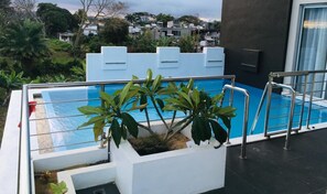 A side view of a modern building with a rooftop pool, surrounded by greenery and overlooking a residential area.