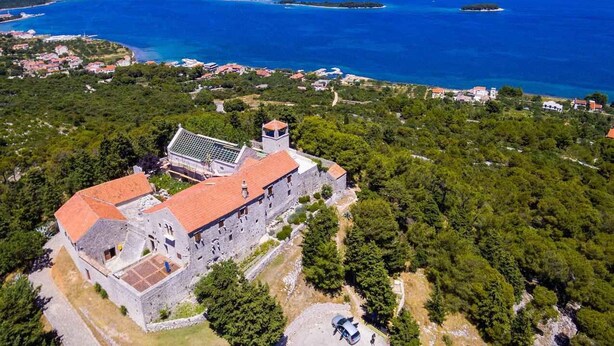 An aerial view of a historic stone building with a red-tiled roof, surrounded by dense greenery and overlooking a large body of water with several islands. - Holiday apartment with air conditioning and sea view (Tkon)