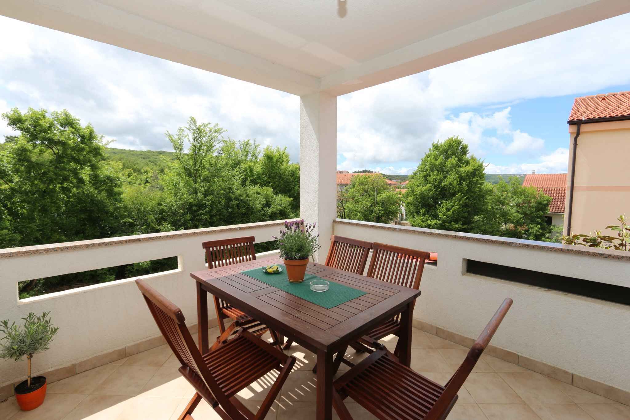 A covered outdoor seating area with a table and chairs, surrounded by a white wall with windows and a view of green trees and buildings.