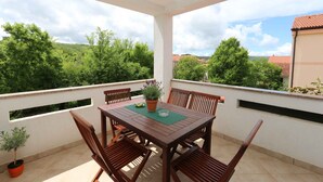A covered outdoor seating area with a table and chairs, surrounded by a white wall with windows and a view of green trees and buildings.