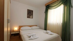 A neatly made bed with white linens and two pillows, a bedside table with a lamp, a framed picture on the wall, and green curtains on the window.