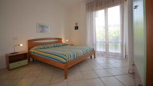 Un dormitorio con cama de madera, mesita de noche, cajón pequeño, luz empotrada en la pared, ventana con cortinas translúcidas y una puerta.