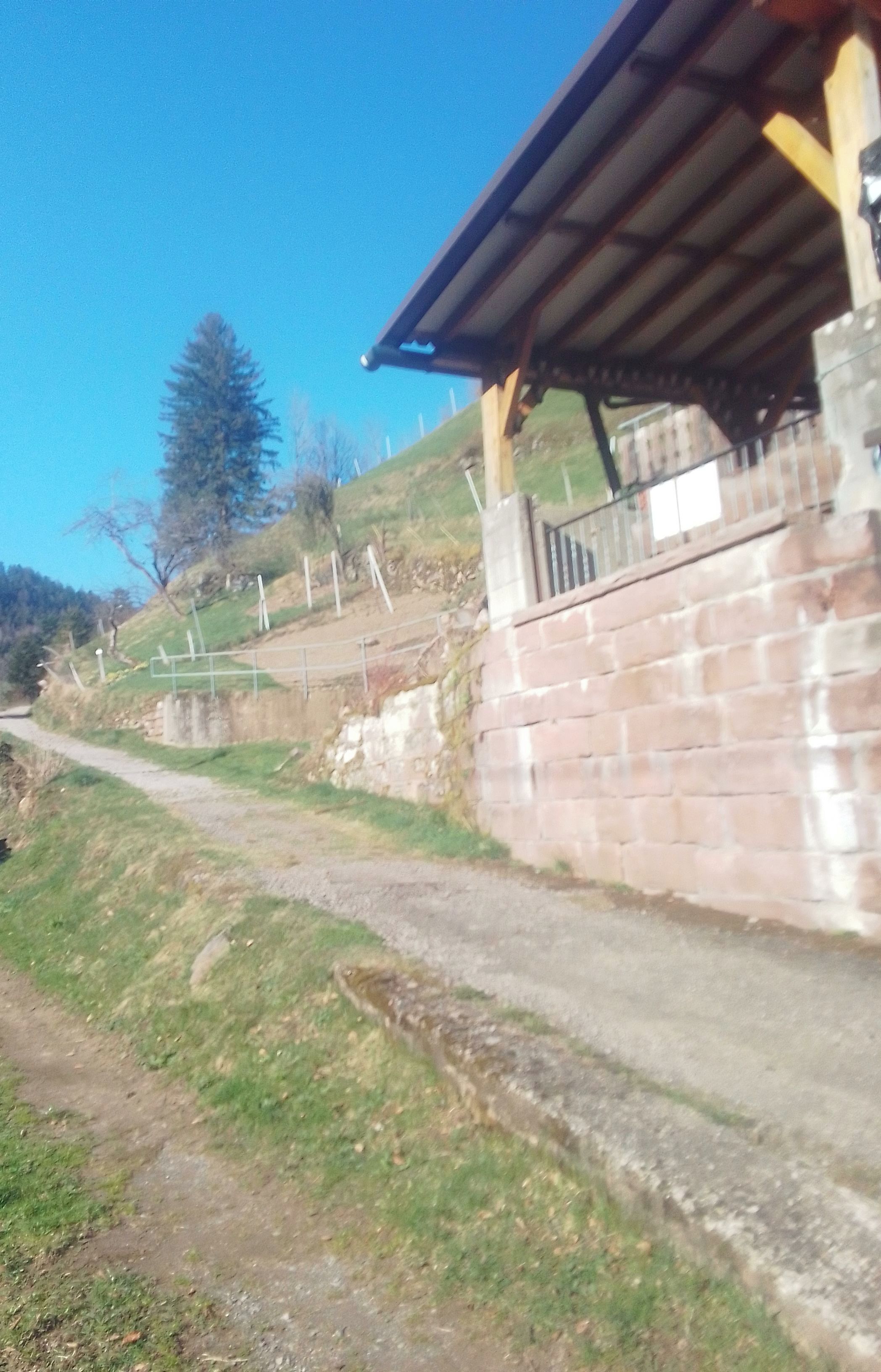 Uma estrada rural, com um muro de tijolos de um lado e uma construção de madeira do outro.