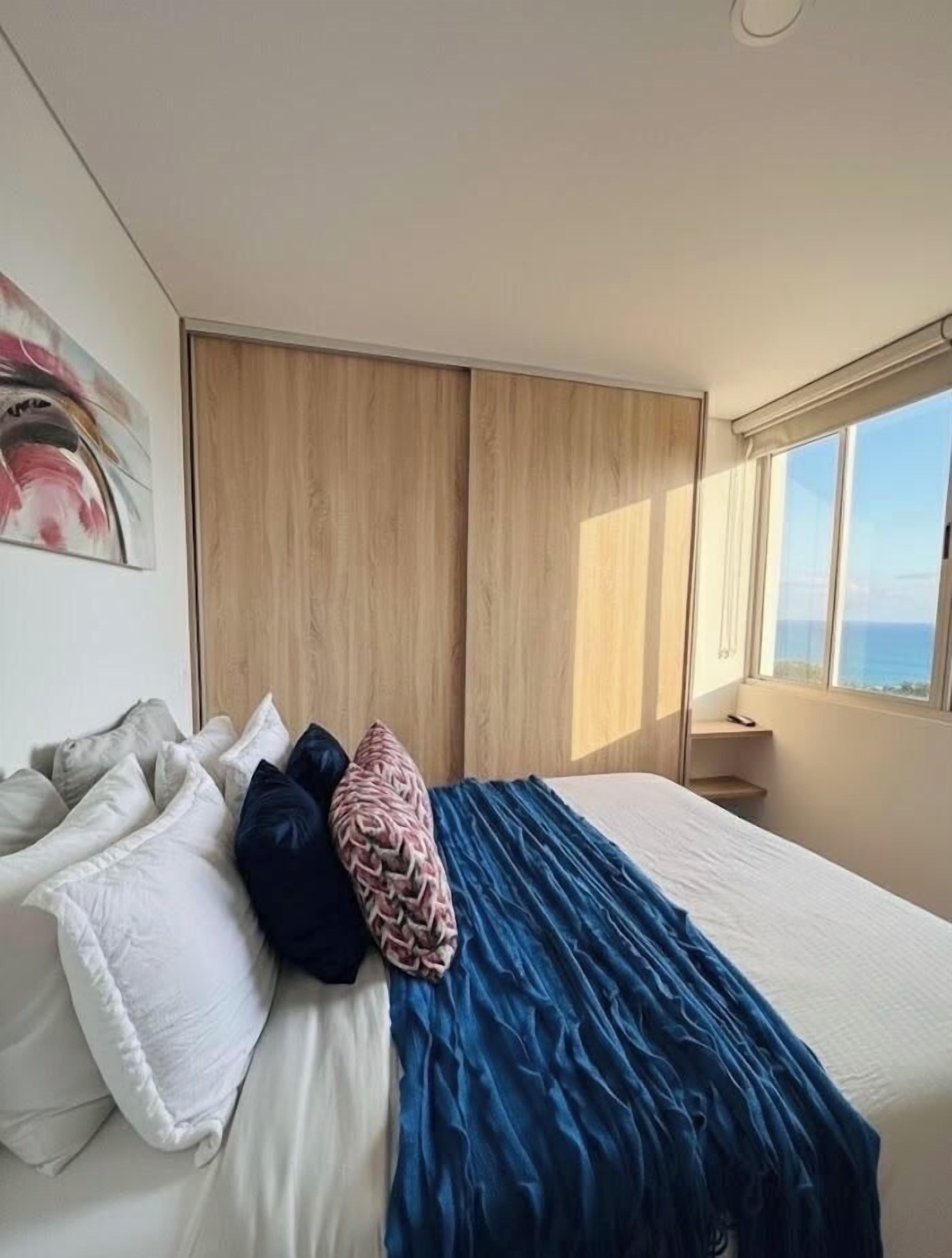 A bedroom with a bed, pillows, a blue blanket, a wooden wardrobe, and a window with a view.