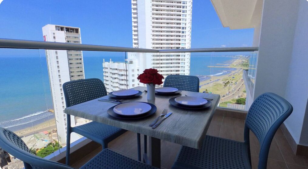 A balcony table with four chairs, a vase with a red flower, and a view of the ocean and cityscape.