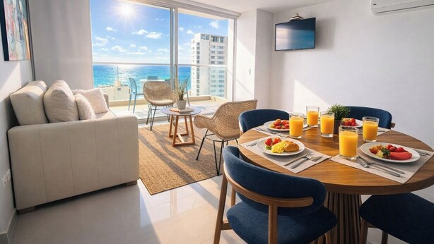 Una sala de estar moderna con un sofá, una mesa de comedor y sillas, un ventanal con vistas al mar y un televisor de pantalla plana montado en la pared.