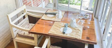 Una mesa de madera con mantel blanco, dos sillas blancas con cojines estampados y un ventanal con vistas a follaje y un muro de ladrillo.