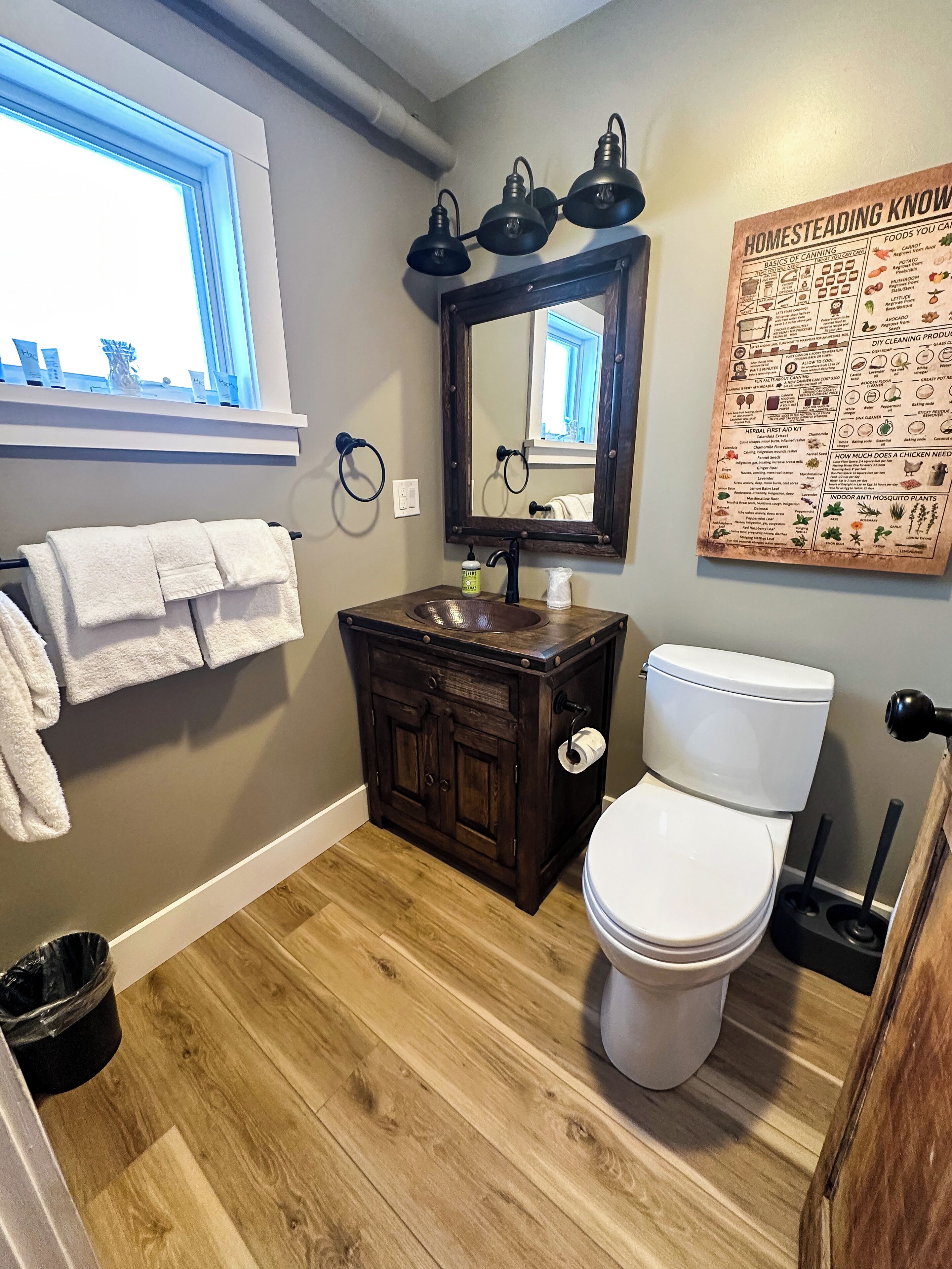 A bathroom with a wooden vanity, a white toilet, a mirror with three lights, and a window.