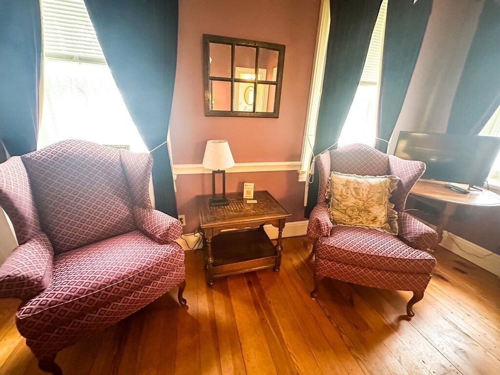 A room with two patterned armchairs, a small table with a lamp, and a window with curtains.
