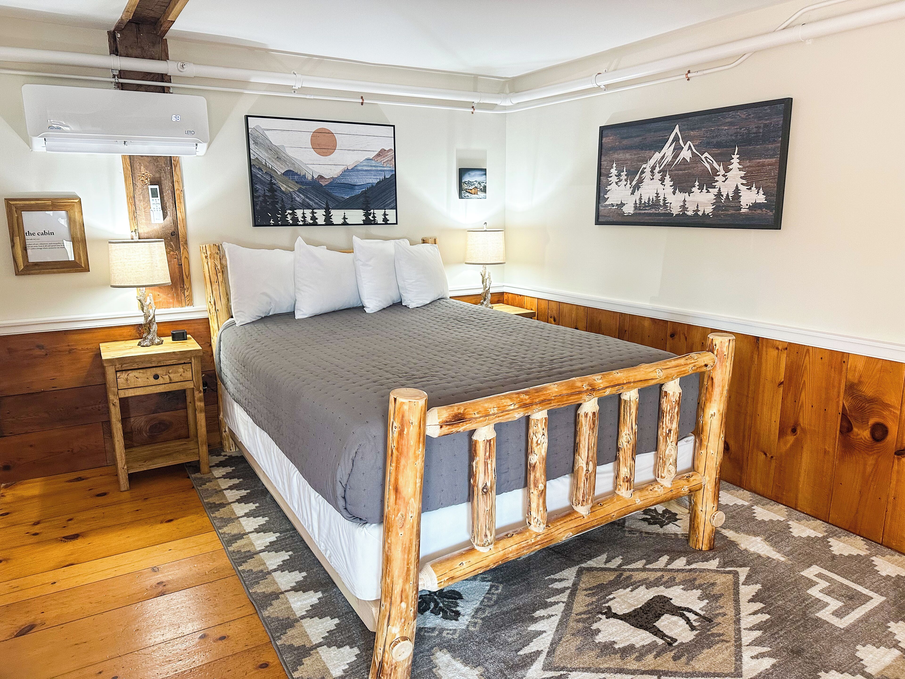 A bedroom with a wooden bed frame, a gray bedspread, a nightstand with a lamp, and a wall-mounted air conditioner.