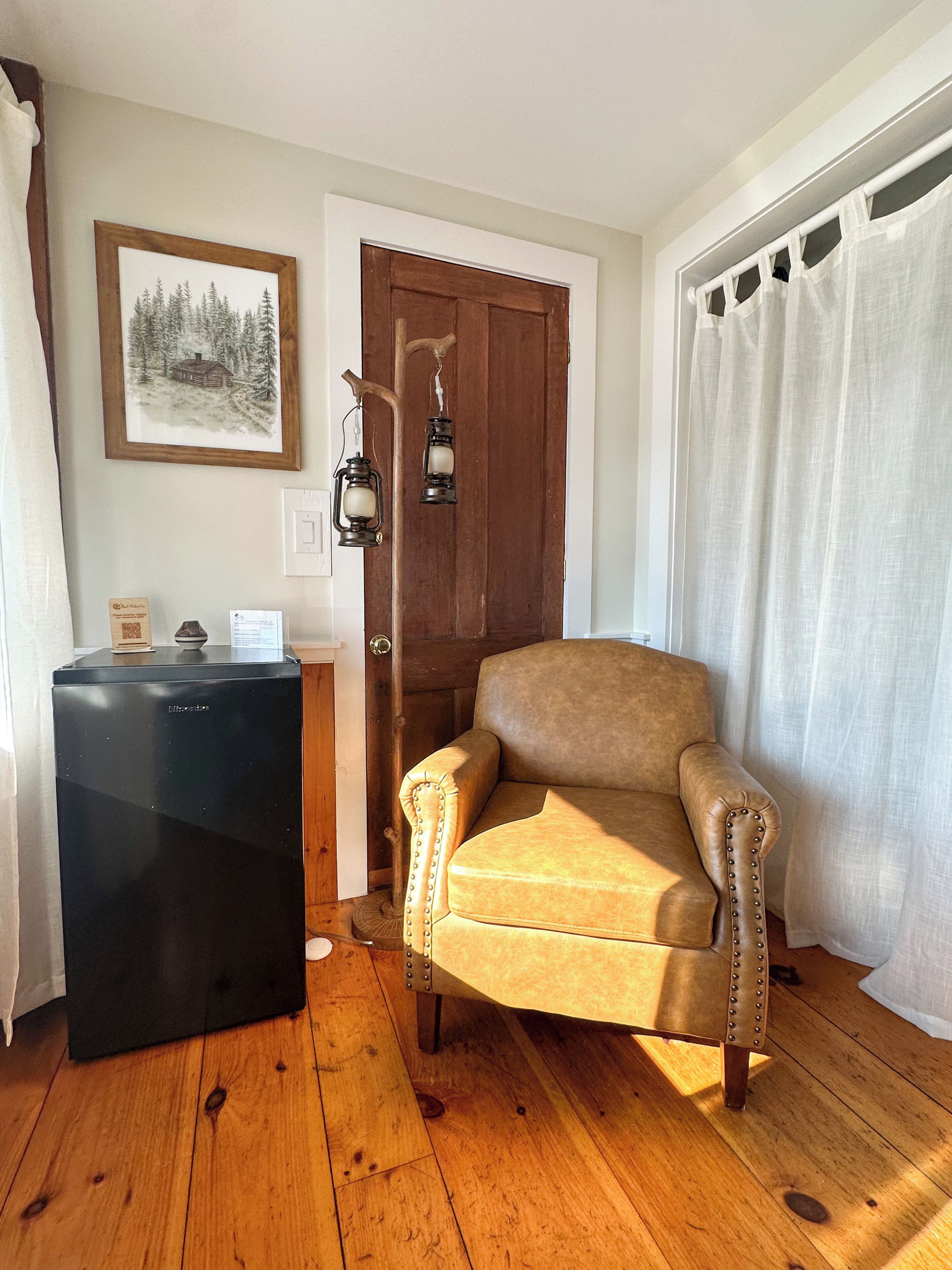 Una habitación con piso de madera, un sillón de cuero marrón, un refrigerador negro, una puerta de madera y una cortina blanca.