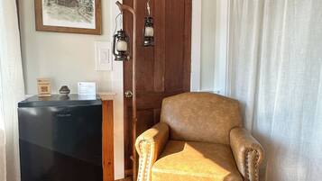 Una habitación con piso de madera, un sillón de cuero marrón, un refrigerador negro, una puerta de madera y una cortina blanca.