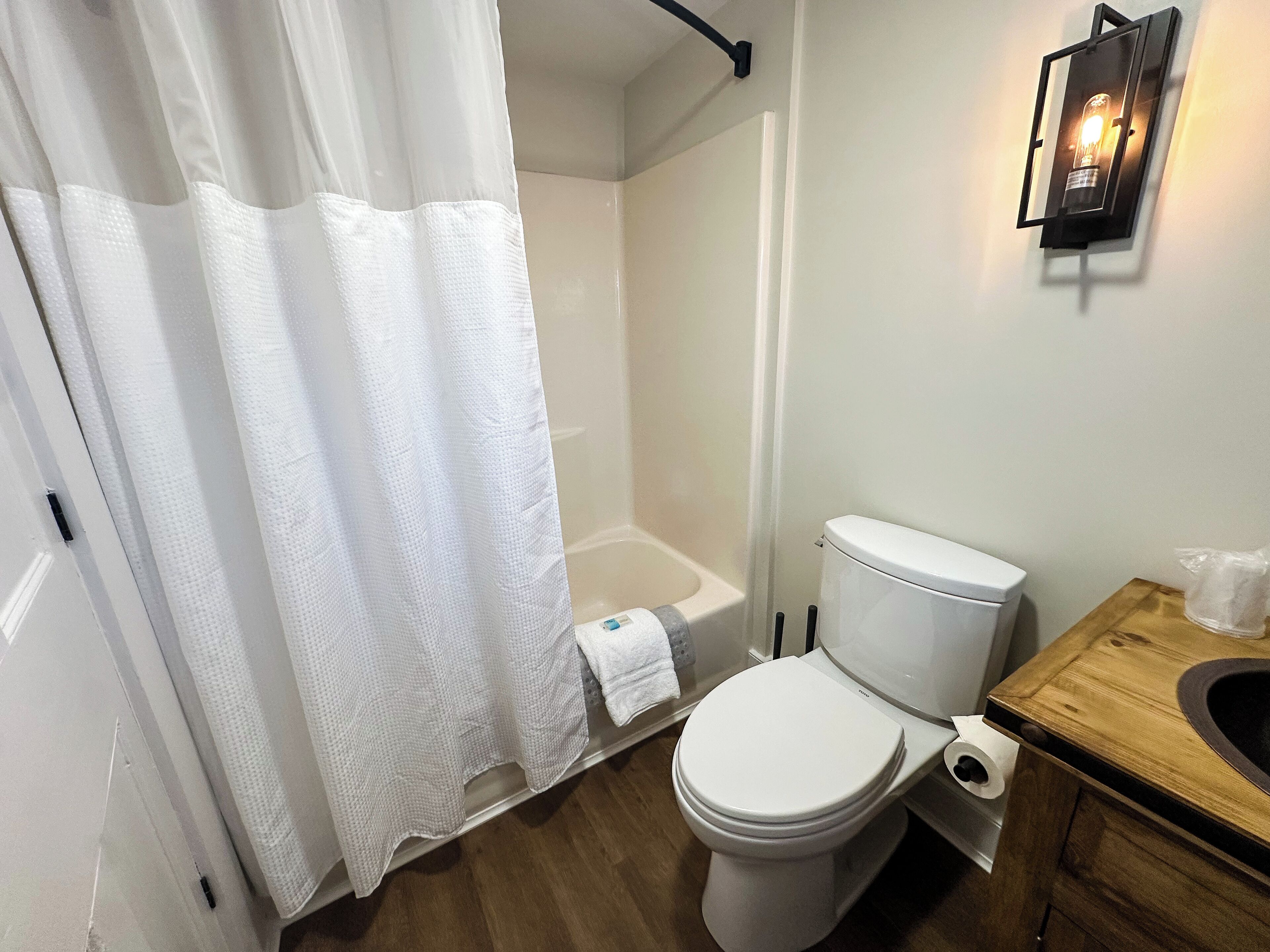 A bathroom with a toilet, a wooden vanity, a shower curtain, and a wall-mounted light fixture.
