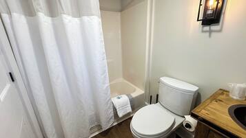 A bathroom with a toilet, a wooden vanity, a shower curtain, and a wall-mounted light fixture.