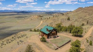 A wooden cabin with a green roof, surrounded by a dirt path and sparse vegetation, set against a backdrop of rolling hills and distant mountains. - 24-0089 Rustic Hartsel Getaway w/ 35-Acre Yard (Fairplay)