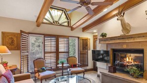 A cozy living room with a fireplace, a ceiling fan, and a large window with shutters.
