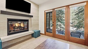 A modern living room with a fireplace, a television mounted on the wall, and a sliding glass door leading to an outdoor area with trees. - The Charter at Beaver Creek E0405 (Beaver Creek)