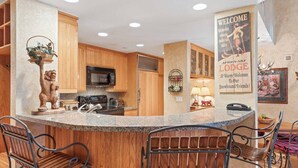 A kitchen with wooden cabinets, a granite countertop, and a bear statue. There are three metal bar stools and a welcome sign. - The Charter at Beaver Creek D4205 (Beaver Creek)