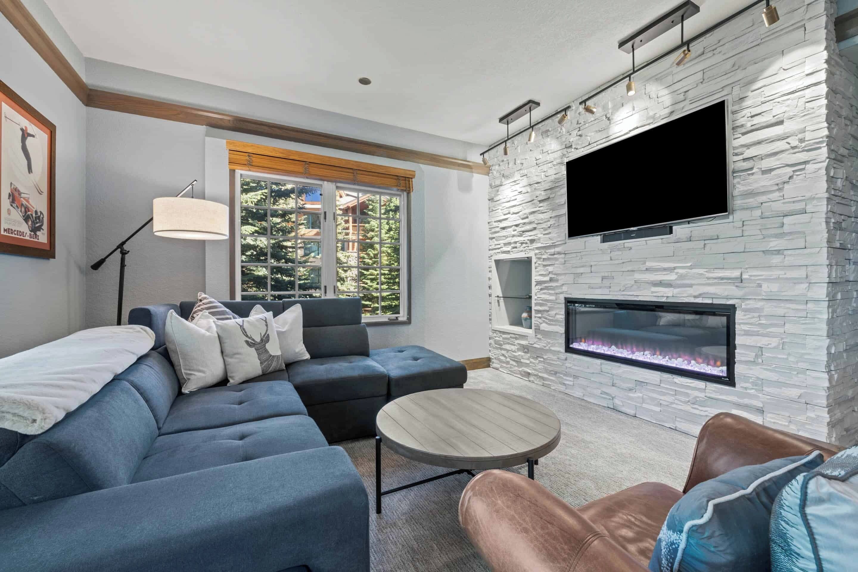A modern living room with a sectional sofa, a coffee table, a fireplace, and a large TV mounted on a stone wall.
