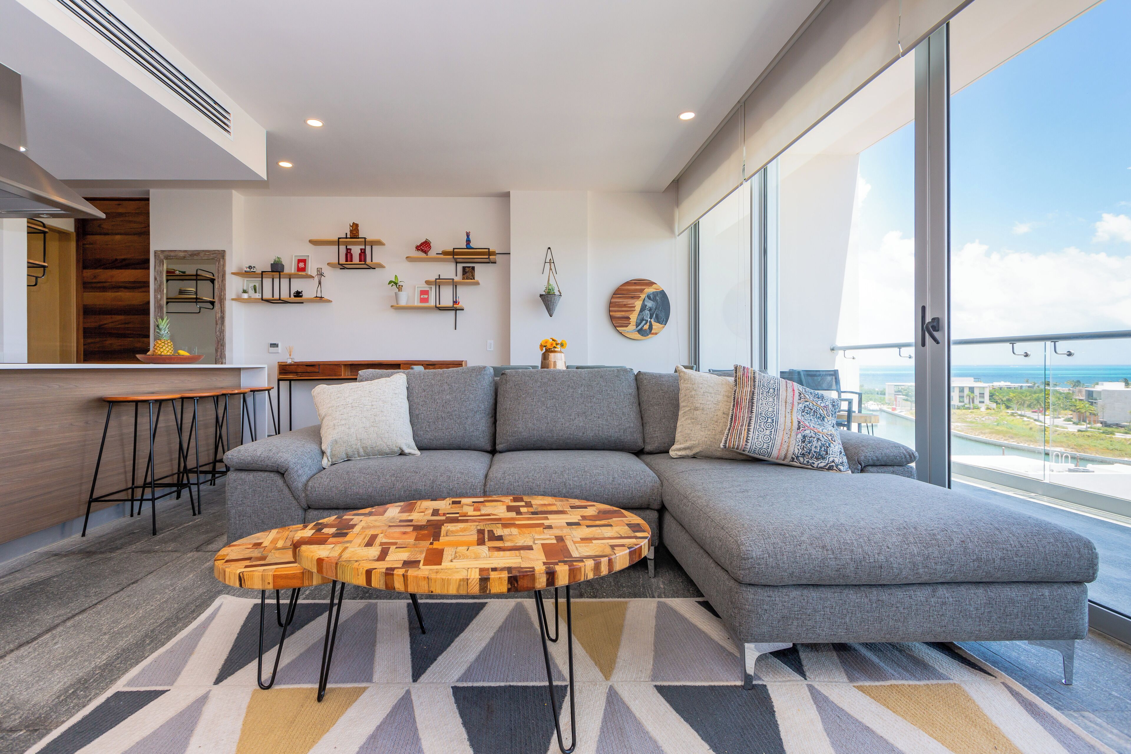 Uma sala de estar moderna com um sofá cinzento, uma mesa de centro de madeira e vista para a cidade através de grandes janelas.