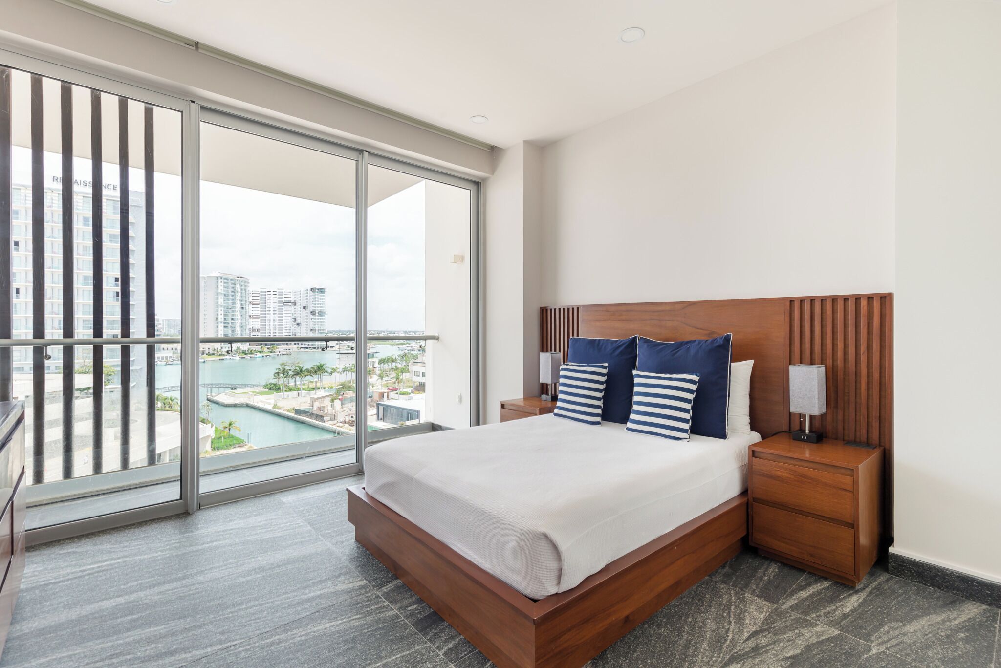 A modern hotel room with a large sliding glass door leading to a balcony, a wooden bed with a headboard, and a nightstand with a lamp.