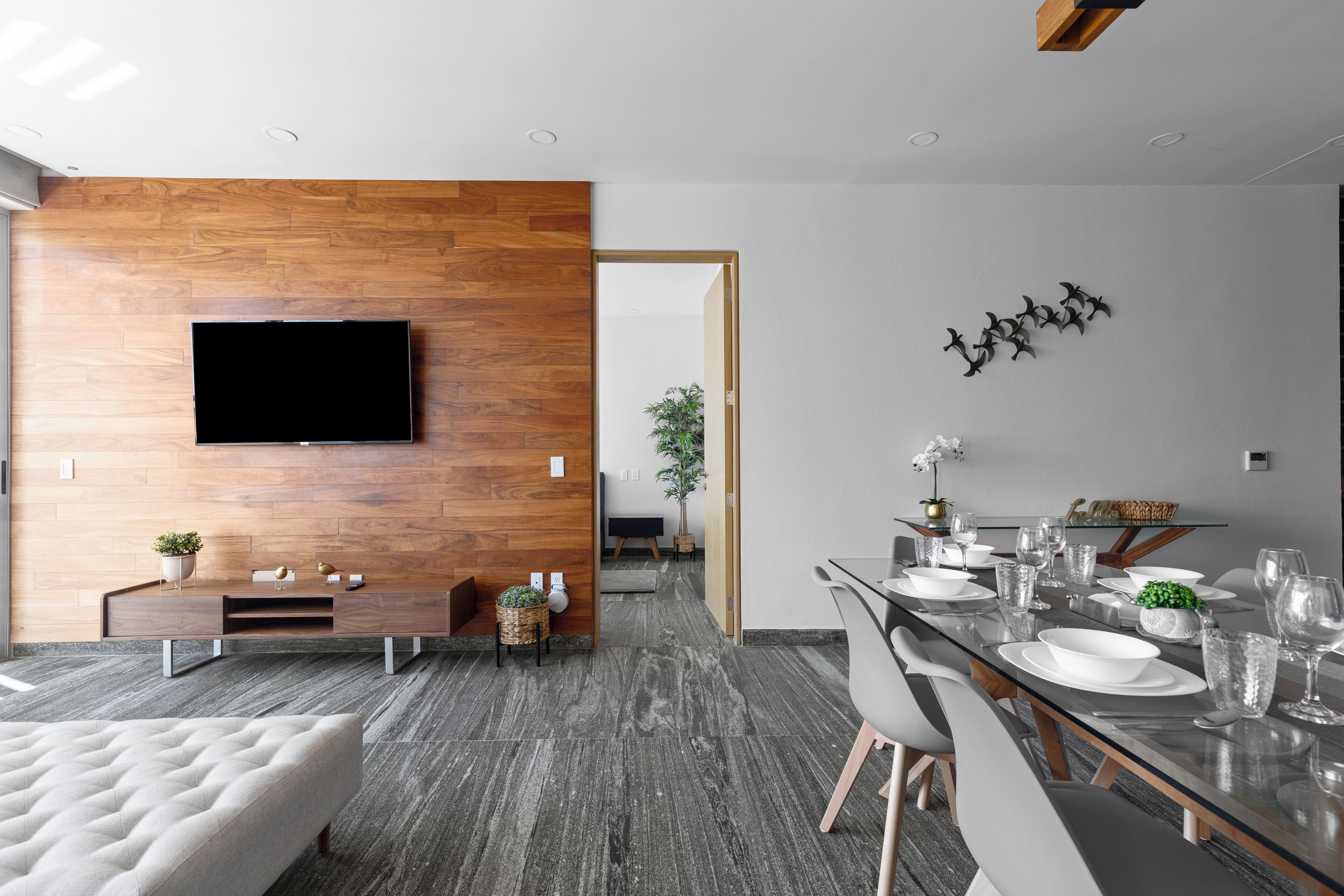 A modern living room with a flat-screen TV mounted on a wooden wall, a dining area with a glass table and chairs, and a grey tiled floor.