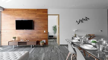 A modern living room with a flat-screen TV mounted on a wooden wall, a dining area with a glass table and chairs, and a grey tiled floor.