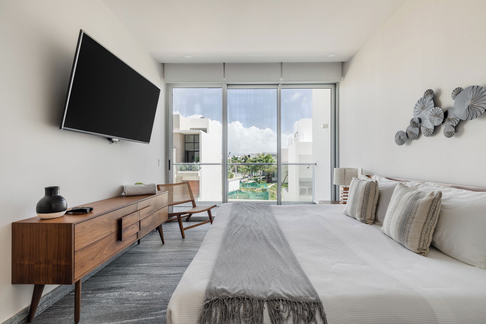 Ein modernes Schlafzimmer mit einem großen Bett, einem flachbildfernseher an der Wand, einer Holzkommode und Blick ins Freie durch eine Glastür.