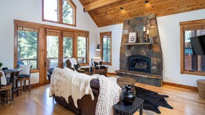 A spacious living room with a stone fireplace, wooden ceiling beams, large windows, and a cozy seating area. - Hot tub! Ski In/Out! Pool Table/PingPong Fireplaces! Mere feet from the Village! (Truckee)