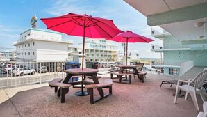 Een balkon met picknicktafels en rode parasols, met uitzicht op een kustgebied met gebouwen en geparkeerde auto's.