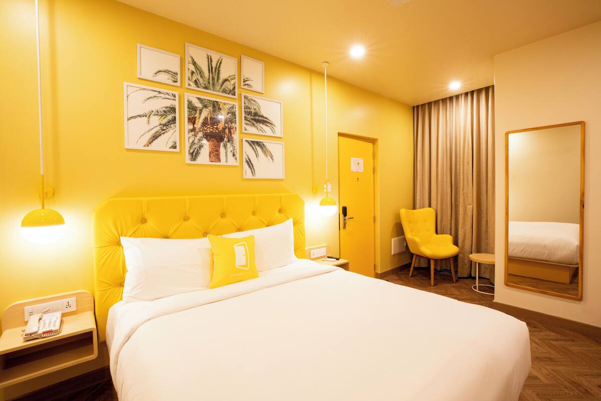 A hotel room with a large bed, a yellow headboard, a bedside table with a phone, a chair, and a mirror.