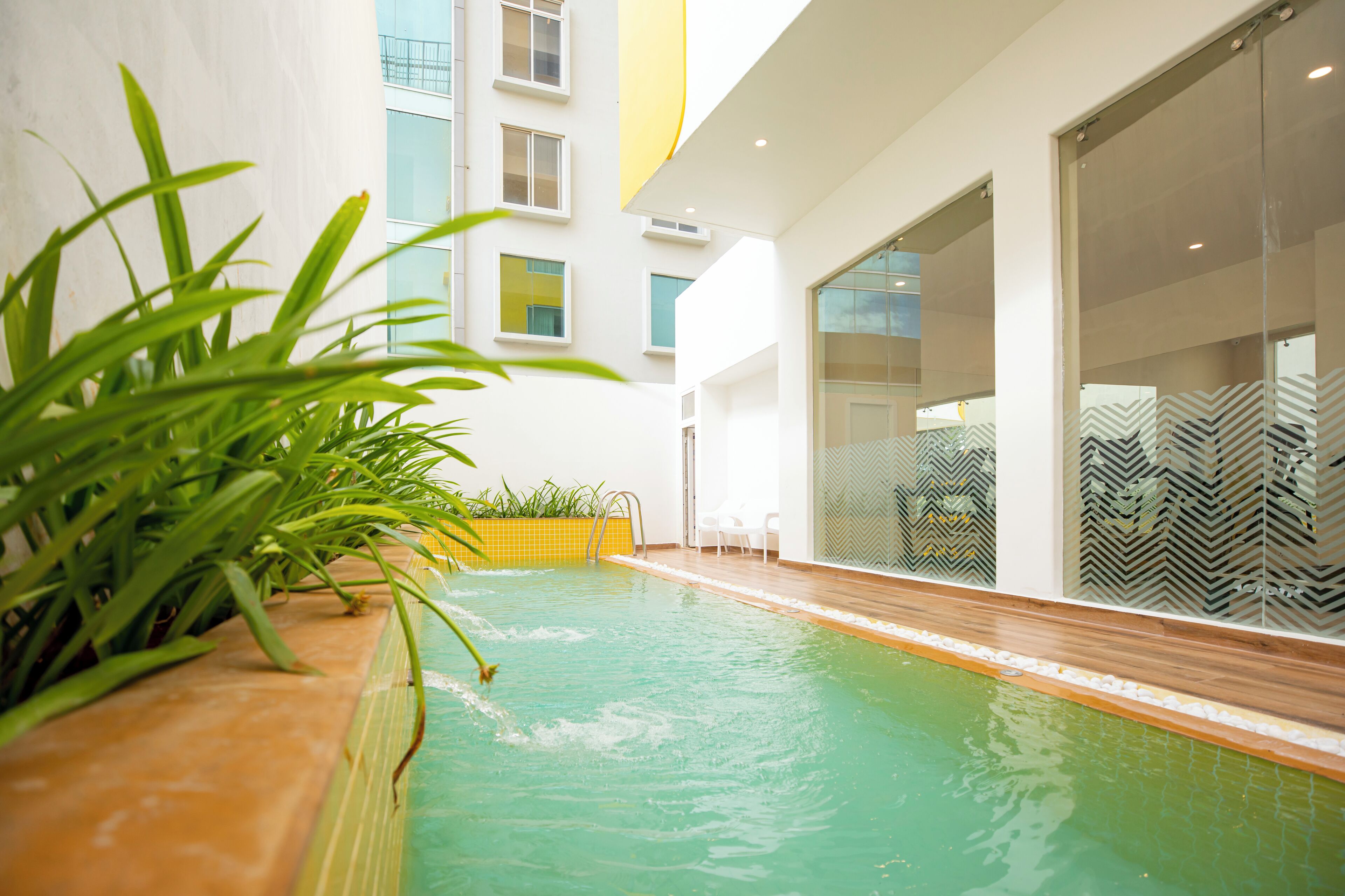 A modern indoor pool area with clear glass walls, a wooden deck, and potted plants.