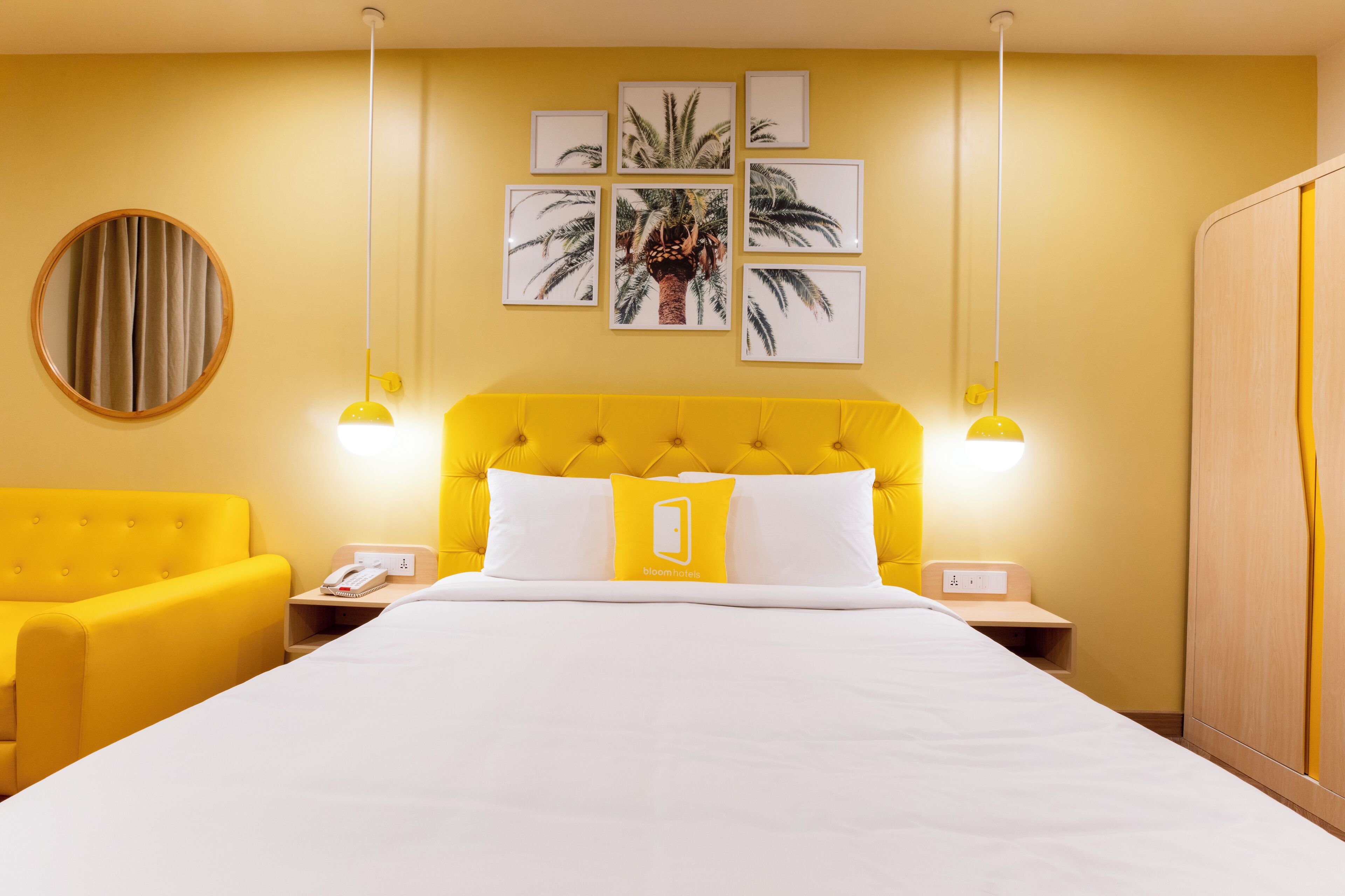 Habitación de hotel con cabecera amarilla, ropa de cama blanca, un espejo redondo y una pared decorada con cuadros.