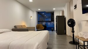 A modern hotel room with a large bed, a desk, a chair, a refrigerator, and a fan. - Urban Loft: Style and Comfort (Medellín)