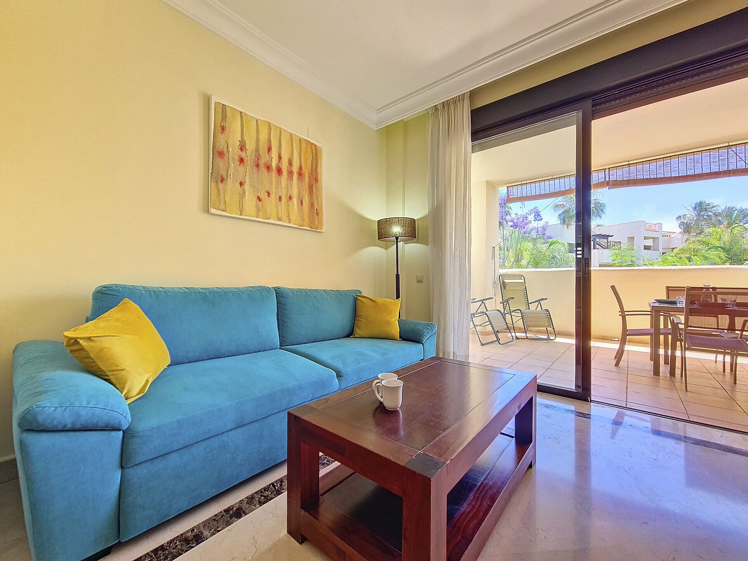 A living room with a blue sofa, yellow cushions, a wooden coffee table, a floor lamp, and a view of the outside balcony area.