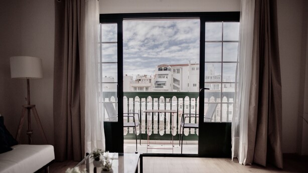 A room with a large window, a sofa, a small table with a plant, and a tripod lamp. - Moderno Apartamento en Puerto de Lacruz (Puerto de la Cruz)