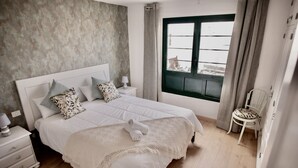 A bedroom with a large bed, a nightstand, a chair, a window with curtains, and a view of the outside. - Moderno Apartamento en Puerto de Lacruz (Puerto de la Cruz)