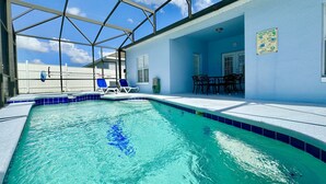 Una piscina con agua cristalina azul, rodeada por una zona cubierta con sillones de descanso y una mesa.