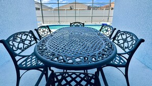 Une table ronde en métal, ornée de motifs complexes, entourée de chaises assorties, le tout placé devant une piscine, avec un ciel dégagé et des bâtiments visibles au loin.