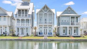 A row of waterfront houses with balconies, a grassy lawn, and a calm water body in the foreground. - Sunkissed Palms 116 NEI (Port Aransas)