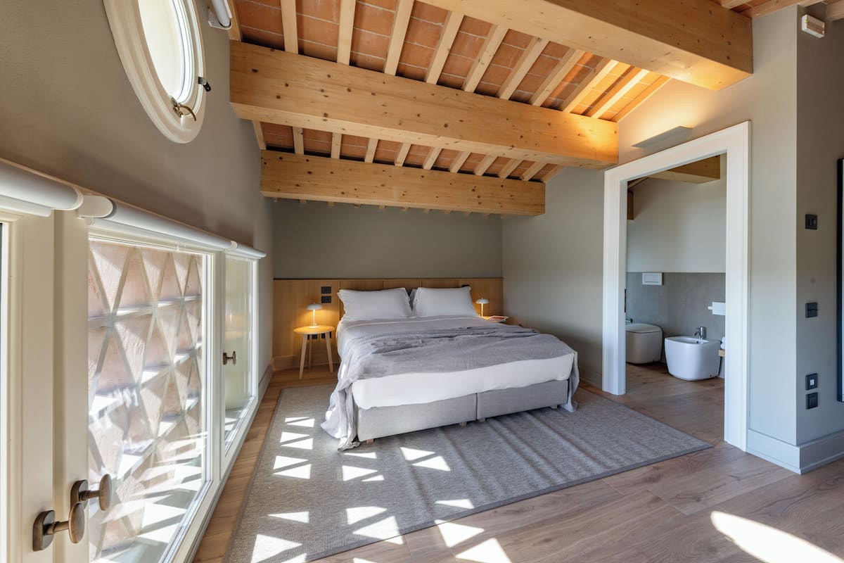 A bedroom with a large bed, wooden ceiling beams, a bathroom, and a view through a glass door.
