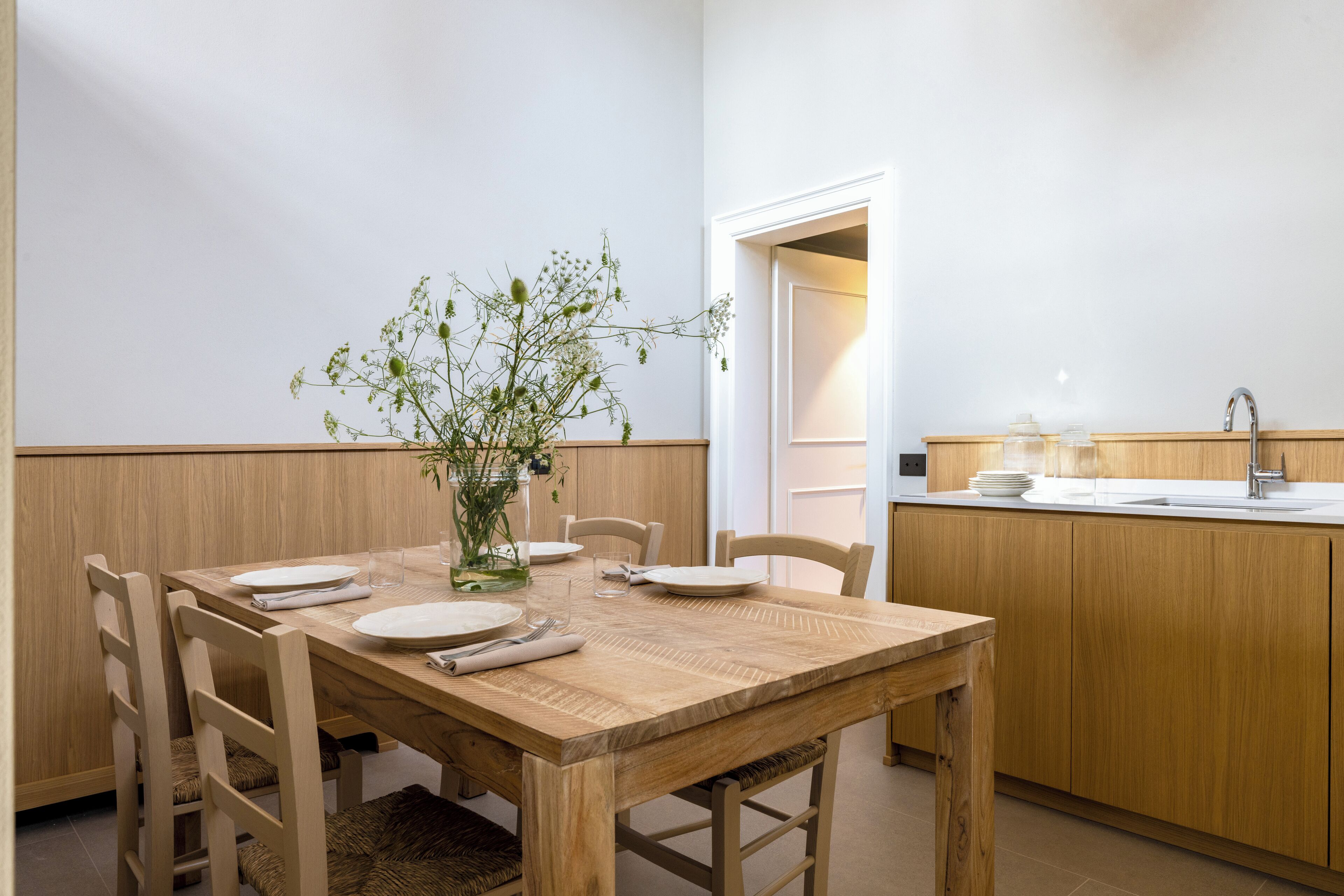 Un comedor con una mesa de madera dispuesta para cuatro, rodeada de sillas, un florero con plantas y una cocina al fondo.
