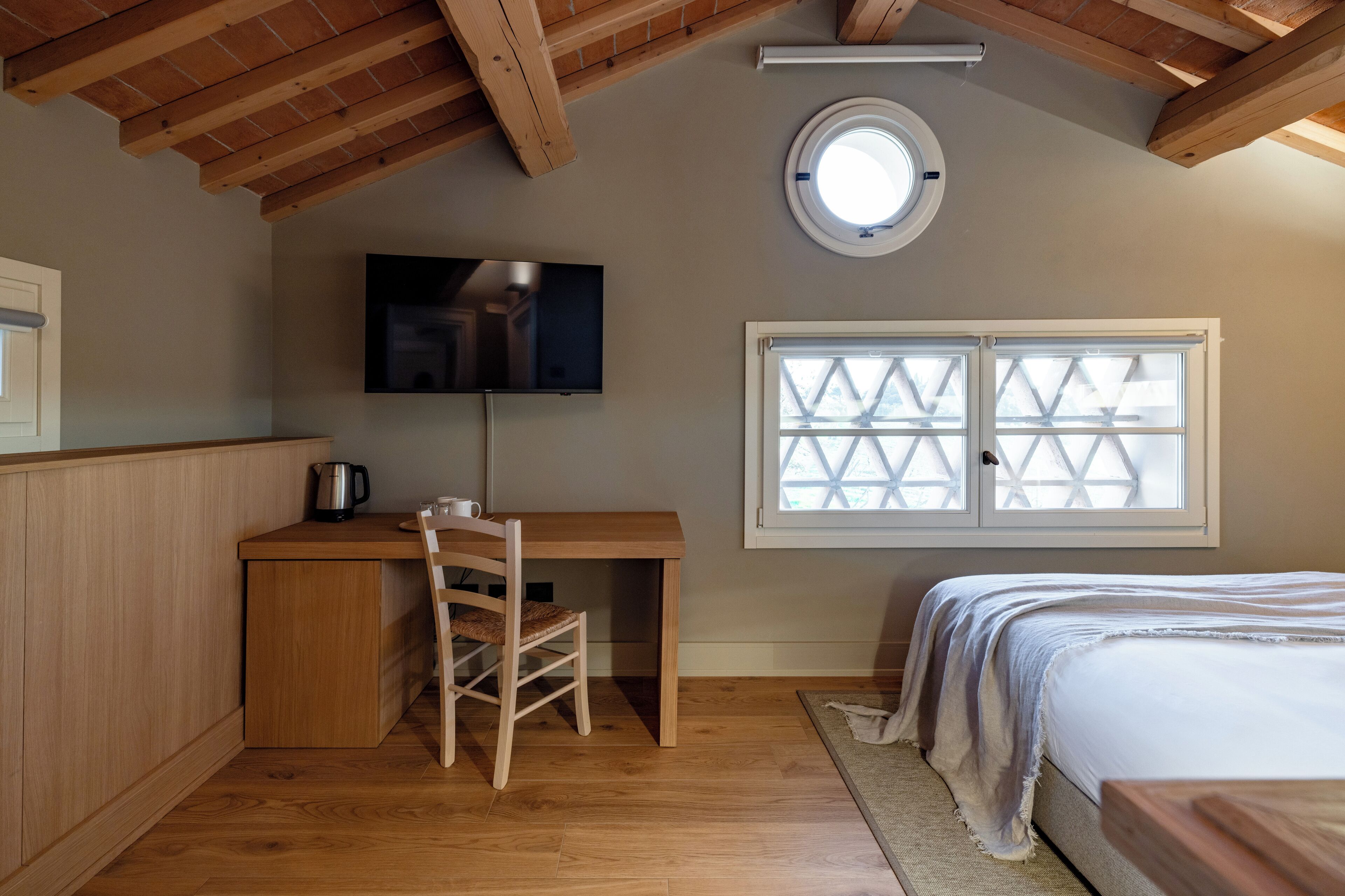 A bedroom with a wooden desk, a chair, a bed, a TV mounted on the wall, and a window with a view of the outside.