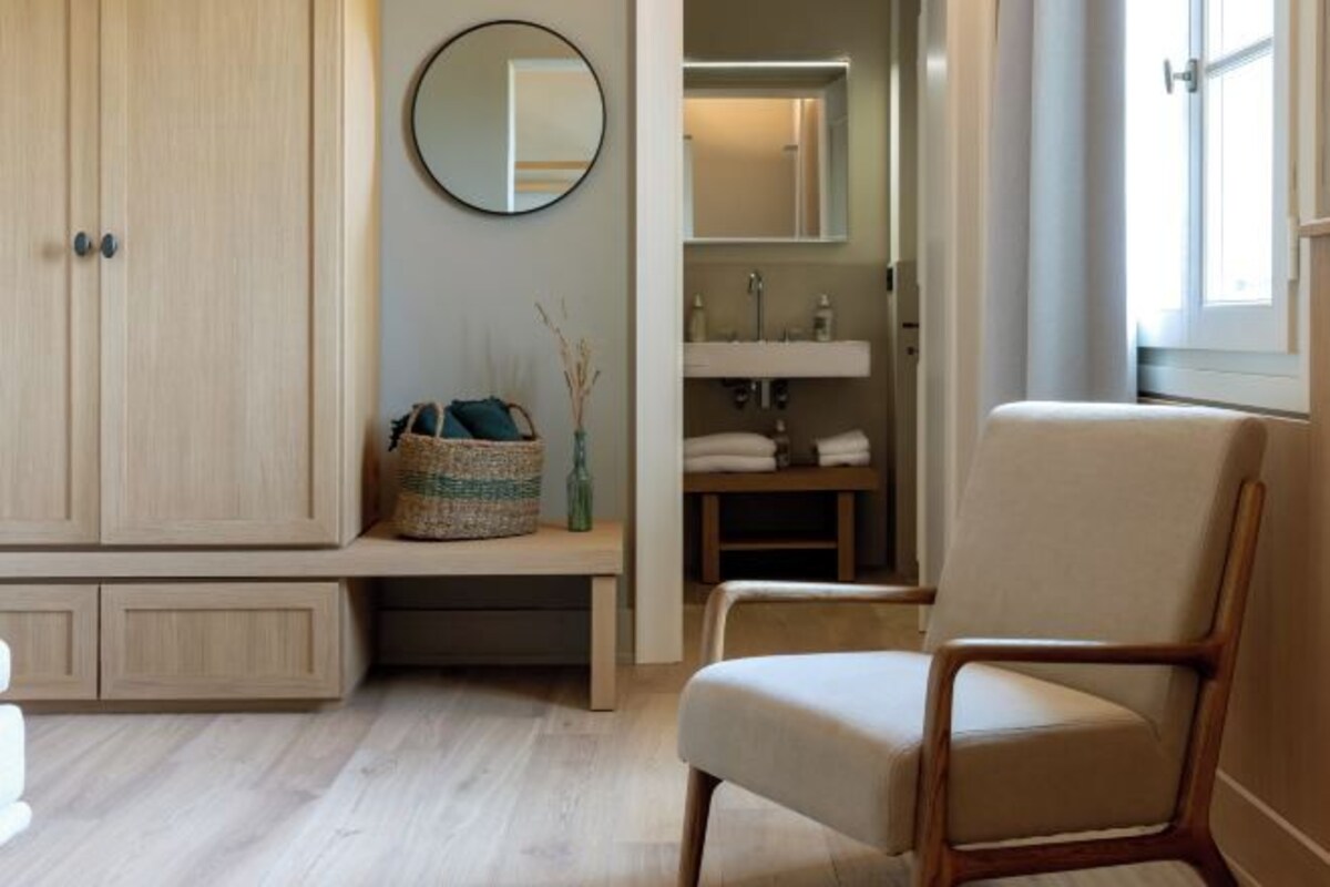 Ein Zimmer mit einem Holzschrank, einem runden Spiegel, einer Bank mit einem gewebten Korb, einem Stuhl und einem Badezimmer mit Waschbecken und Spiegel.