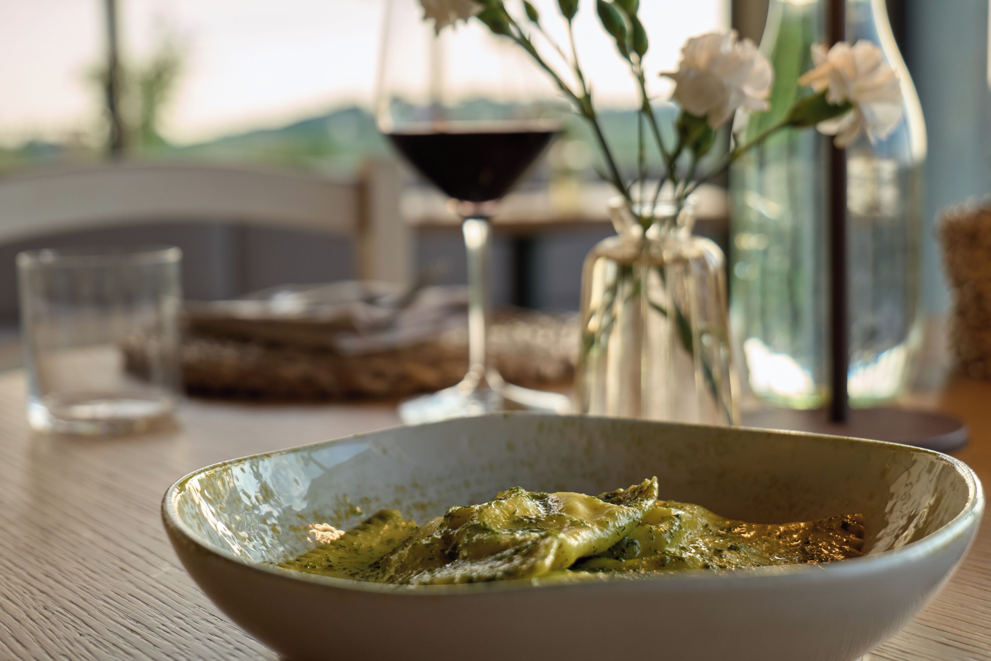 Un tavolo da pranzo con un recipiente pieno di cibo, un bicchiere di vino e un vaso con fiori.